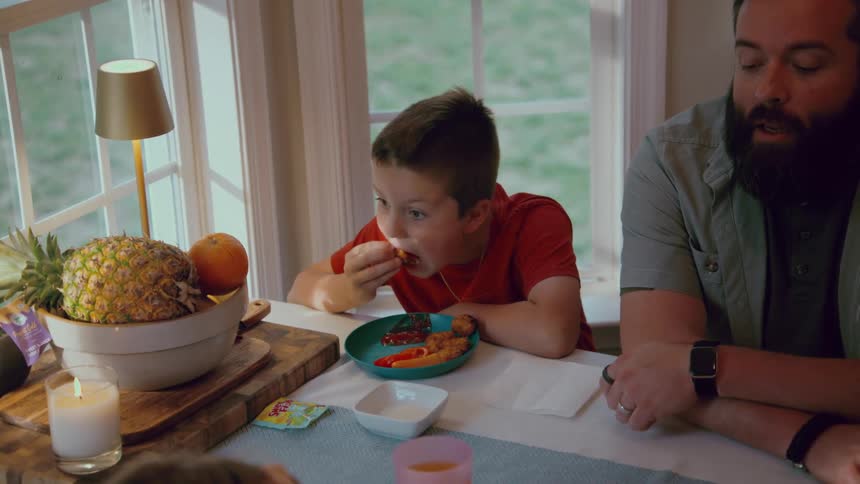 A young boy with a mohawk haircut eats chicken nuggets in slow motion while sitting at the dinner table with his father