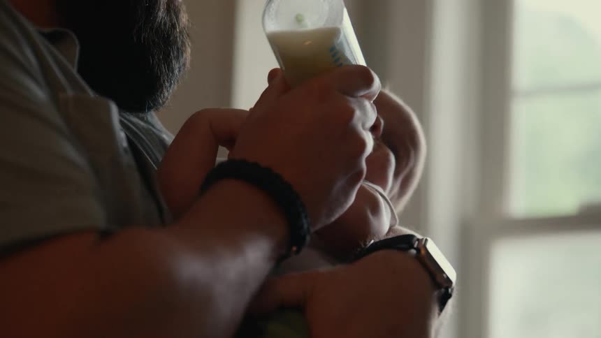 Close up of a father grabbing a baby bottle from the counter and then feeding his newborn child in slow motion