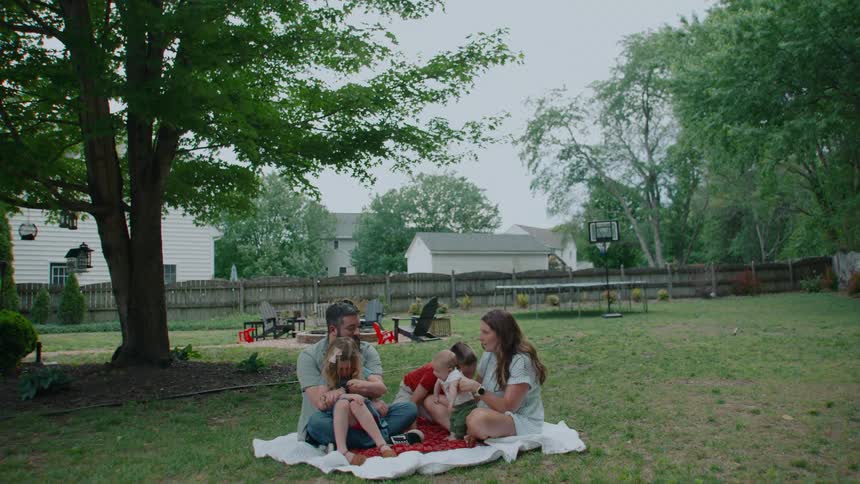 A tilt-down shot reveals a happy family of five enjoying a picnic on a blanket in their spacious suburban backyard