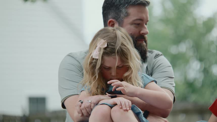 A bearded father watches lovingly as his daughter, sitting on his lap, explores a small creature in her hands outdoors