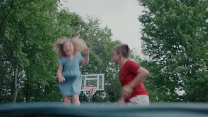 A low-angle slow-motion shot of a brother and sister joyfully jumping on a trampoline in a backyard surrounded by trees