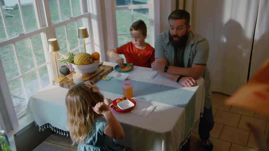 A father tells a story to his son and daughter as they eat breakfast together at a sunny kitchen table