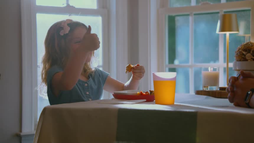 A cute young girl with curly hair eats her dinner at the table and gives a happy thumbs up in slow motion