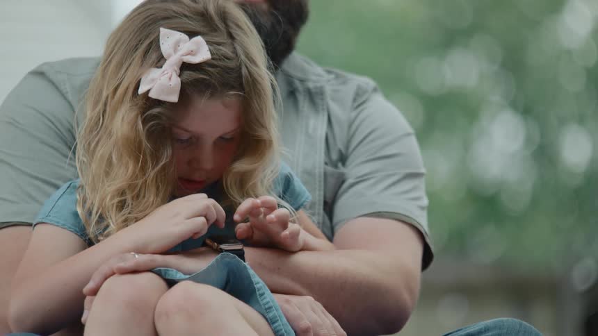 A curious young girl with a pink bow in her hair sits on her father's lap and plays with his smartwatch