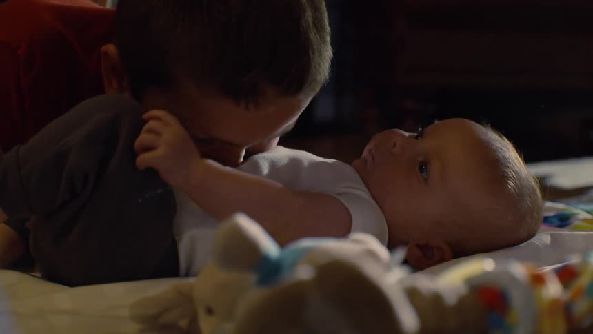 A young boy leans down and lovingly gives his baby sibling a raspberry kiss on the belly as the infant lays on the floor