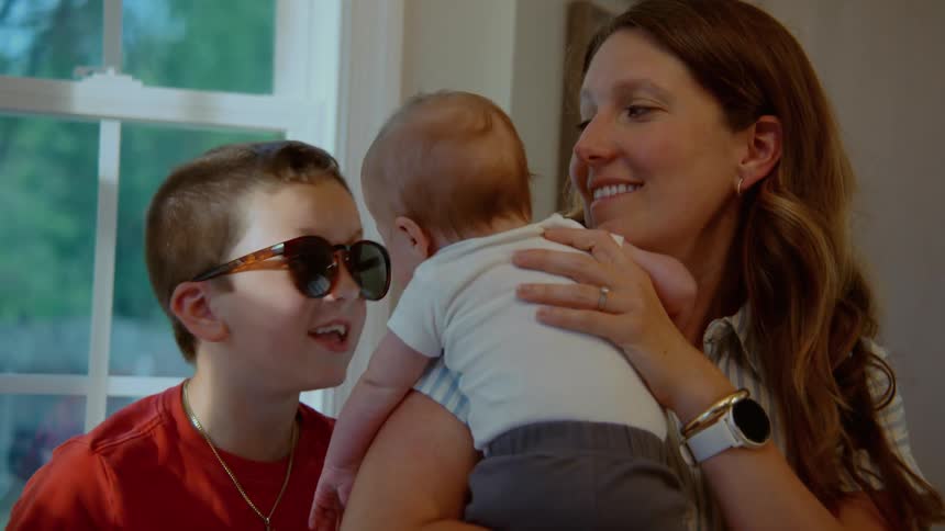 A smiling mother holds her baby while her older son wearing sunglasses talks lovingly to his new sibling in their sunlit home