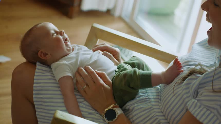 A sweet newborn baby lies in its mother's lap, smiling and laughing during a happy, intimate moment at home