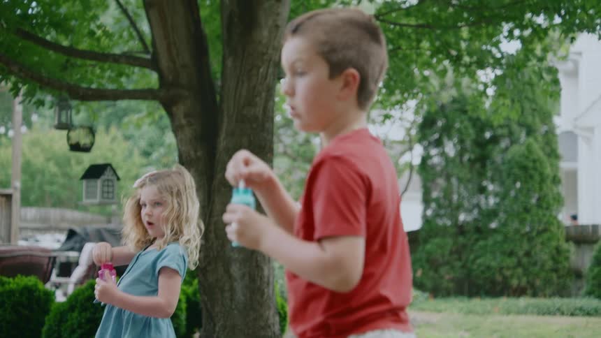 A young boy in a red shirt blows bubbles in slow motion as the camera pulls back to reveal his little sister in the backyard
