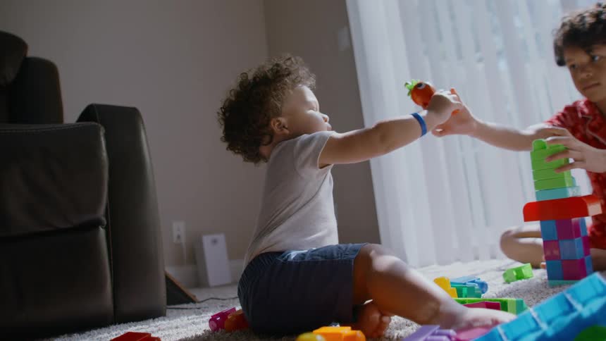 Slow motion footage shows a toddler sharing a toy with his older brother as they play with colorful blocks on the floor