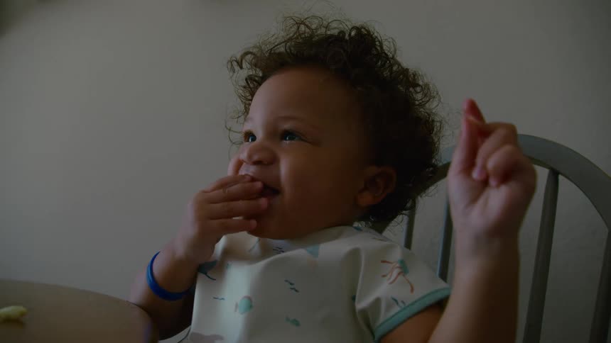 An adorable toddler with curly hair sits in a high chair and laughs happily while eating a snack