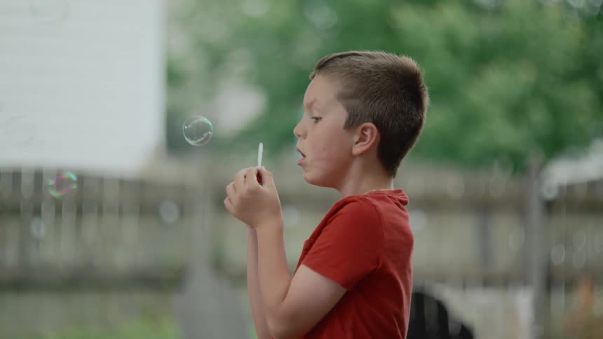 A young boy in a red shirt blows soap bubbles from a wand in his backyard in slow motion