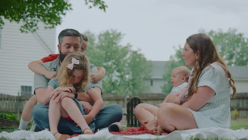 A young boy surprises his father with a hug while the family relaxes together on a blanket in their backyard