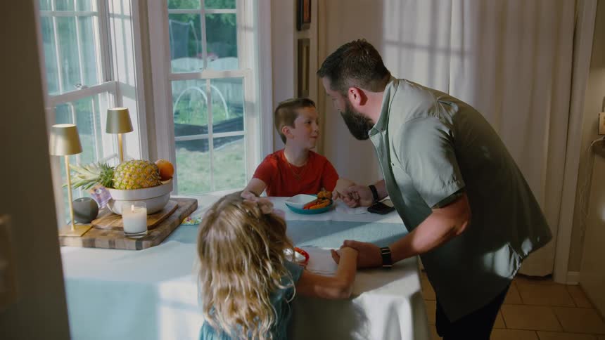 A father and his two children hold hands and pray together before their meal in a sunny dining room