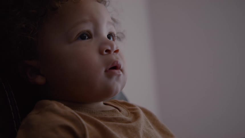 An adorable toddler with curly hair sits mesmerized watching a screen, their face illuminated by the light