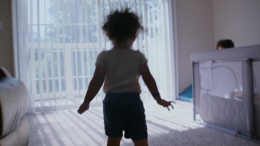 A cute toddler with curly hair happily walks and dances in a sunlit playroom while his brother watches