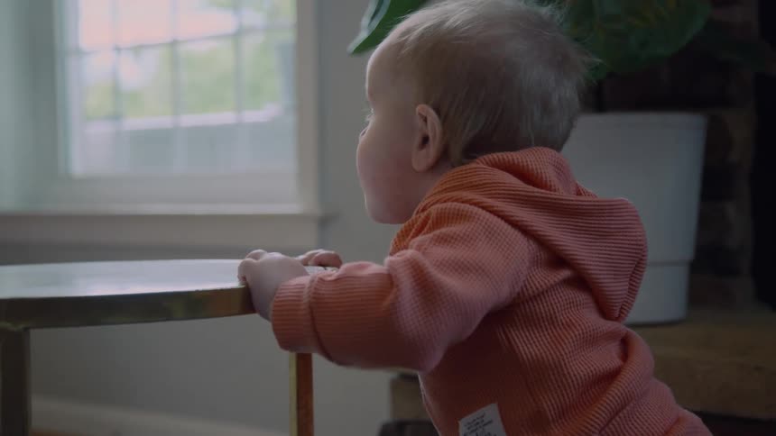 A teething baby in an orange hooded onesie chews on the edge of a metal side table in a naturally lit room