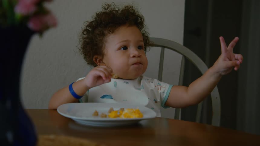 A charming toddler with curly hair sits at a table eating breakfast and playfully raises their hand in a peace sign gesture