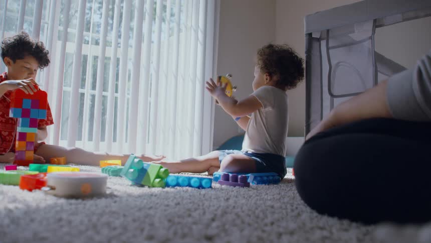 Two young siblings play together with colorful toys on a soft carpet in a sunlit room while their parent watches