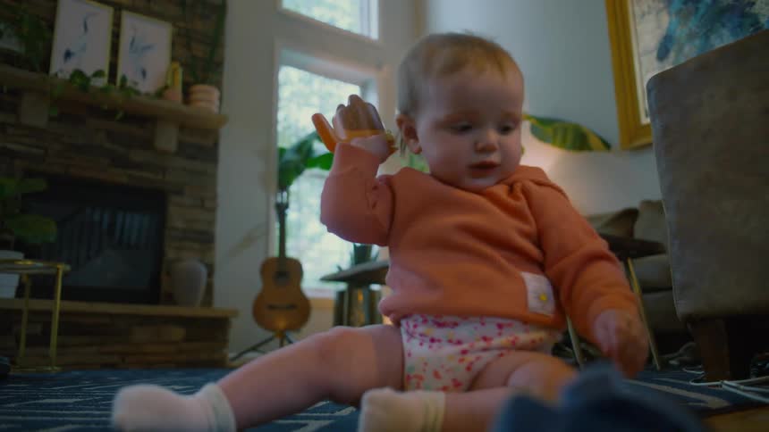 A cute baby wearing an orange hoodie sits on a patterned rug and plays with a soft toy in a cozy living room