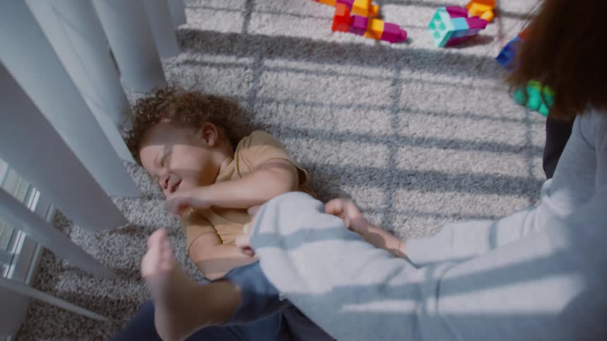 A mother playfully tickles her laughing son on the floor with shadows from the window blinds