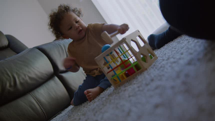 A cute toddler with curly hair energetically hits a colorful wooden toy with a mallet while playing on the living room floor