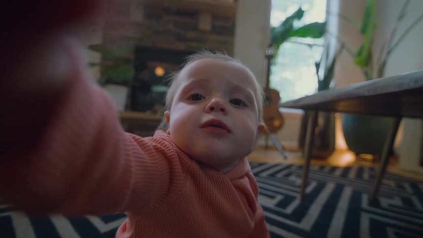 A curious baby reaches for and touches the camera lens from a personal point of view in a cozy living room