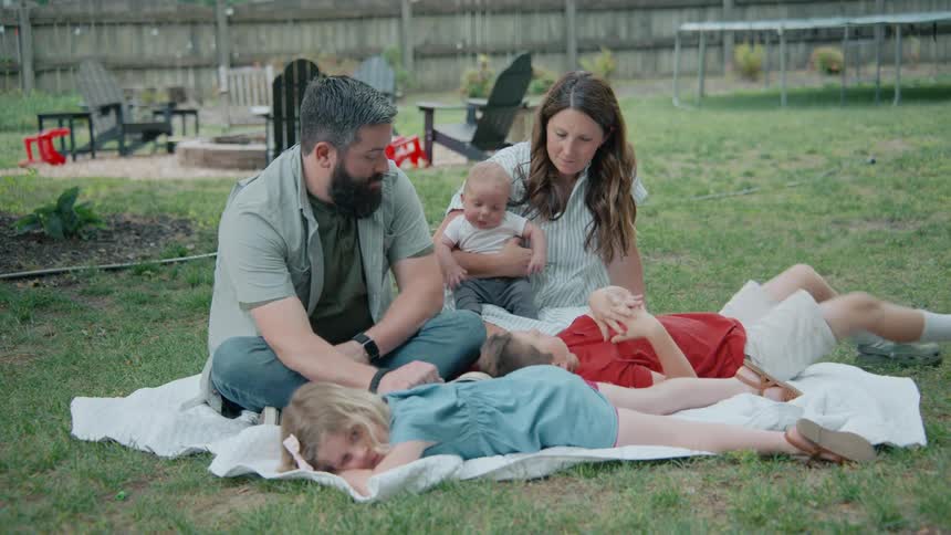 A family of five with a mother, father, two children, and a baby relax together on a blanket in their backyard