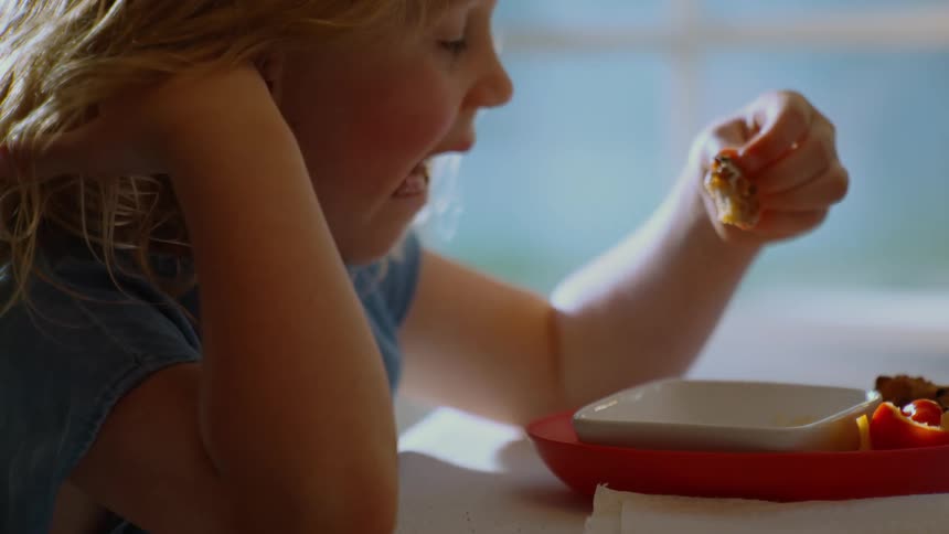 Slow motion profile shot of a little girl with blonde curly hair enjoying a meal by a window