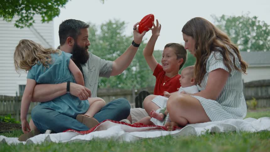 A happy family of five with a newborn baby relaxes on a blanket in their backyard while the older brother plays with a toy