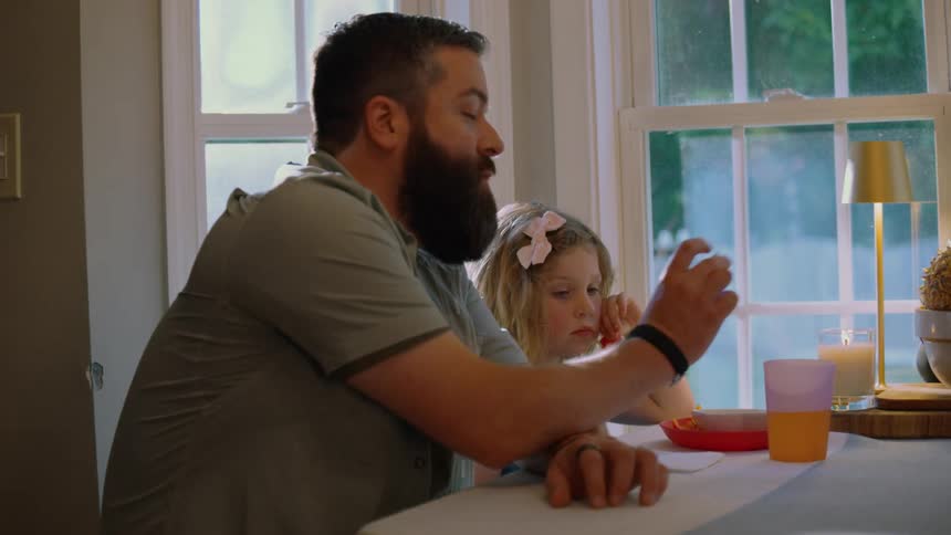A father with a beard and his young daughter sit at a dining table by a window, eating and sharing a quiet family meal together