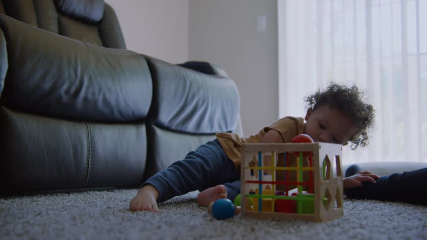 A curious curly-haired toddler sits on the carpet and intently plays with a colorful wooden activity cube in slow motion