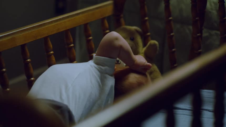 A restless newborn baby lies awake and starts to fuss in a dark wooden crib at night with a stuffed lamb and teddy bear