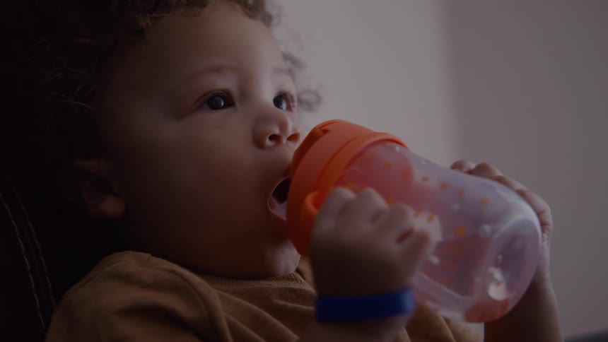 A close-up of a cute toddler with curly hair reaching for and drinking from a sippy cup