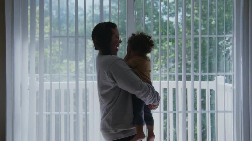A smiling mother holds her toddler in her arms in front of a brightly lit window, sharing a tender moment together