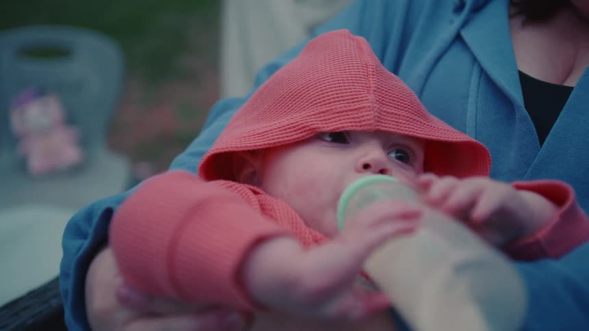 A newborn baby in a pink hooded sweater drinks from a bottle while being held in their mother's arms outdoors