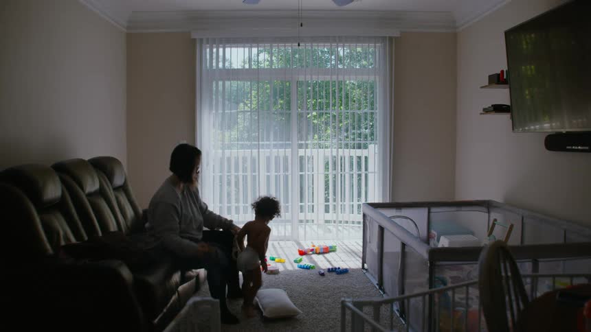 A mother helps her toddler take off his shirt in the living room and watches him play