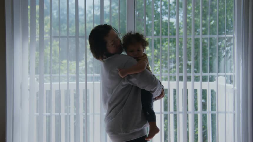 A mother lovingly holds her young child, swaying gently in front of a bright window at home