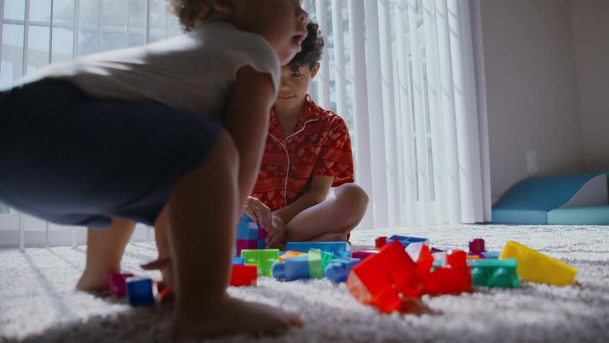 Two young brothers play together with colorful building blocks on a plush carpet in a bright, sunlit room
