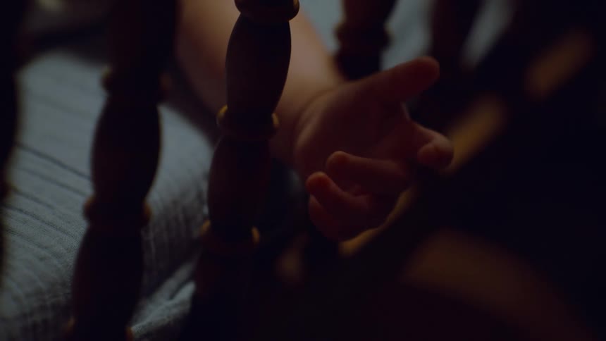 A close-up shot of a baby's tiny hand moving and grasping a wooden crib slat in a dimly lit room