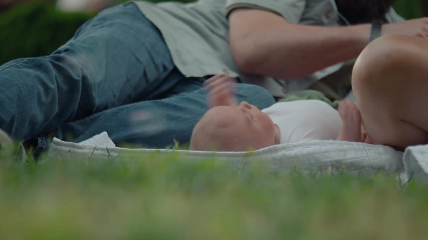 Parents gently caress their newborn baby resting on a blanket in a park as bubbles float past