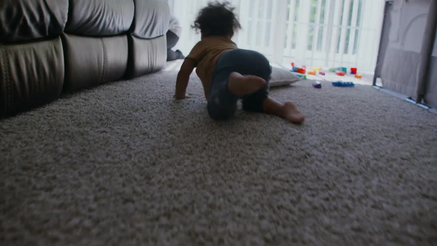 Slow motion, low-angle shot follows a curious toddler as he crawls across a carpeted living room to his toys