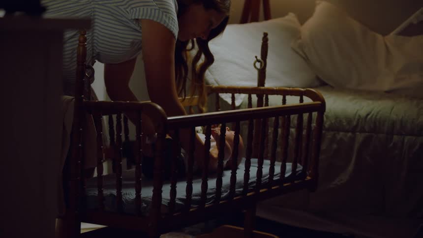 A mother gently puts her newborn baby to sleep in a wooden bassinet in a dimly lit bedroom at night