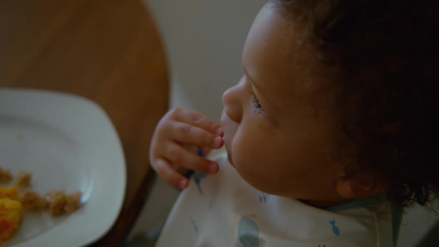 A close-up slow-motion view shows a curly-haired toddler carefully picking up and eating a piece of bread