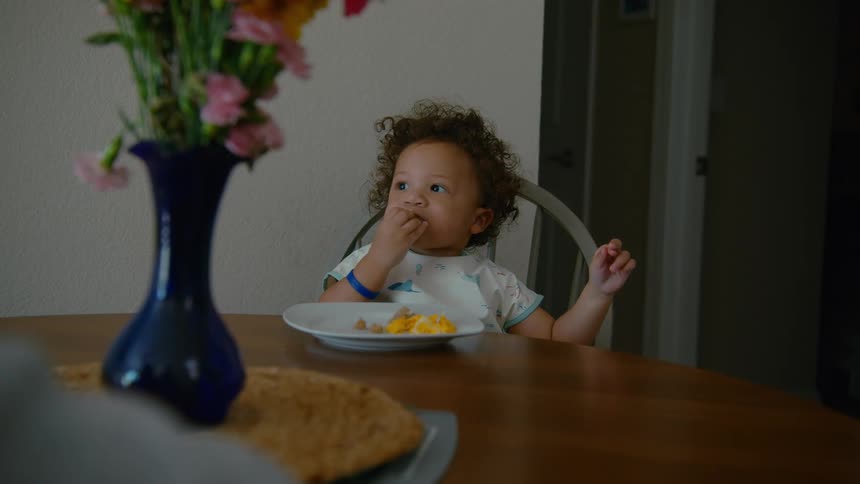 An adorable curly-haired toddler with a bib eats breakfast with their hands while sitting at a dining table