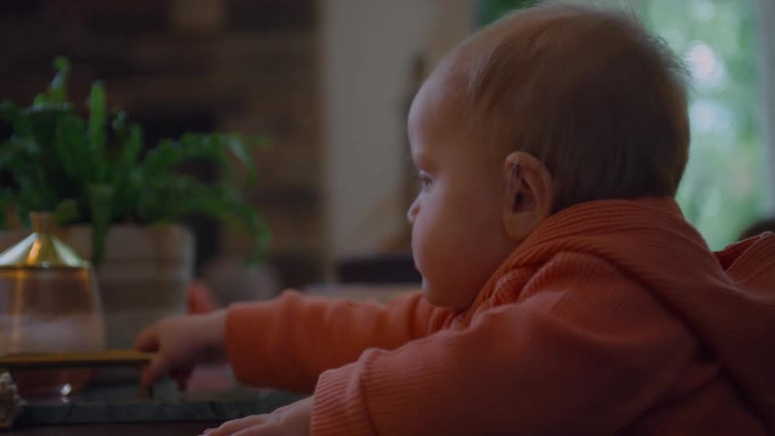A curious baby in an orange hoodie sits at a table, looking around and touching the surface