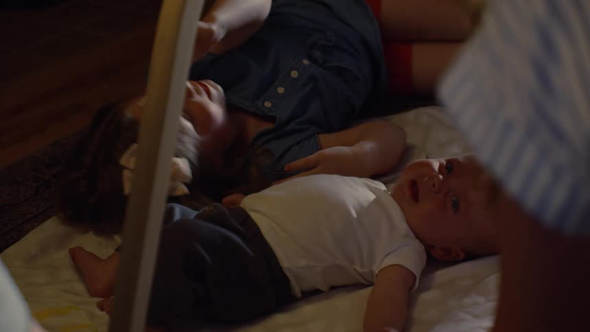 A little girl lies on the floor next to her crying baby sibling while their mother tries to comfort them
