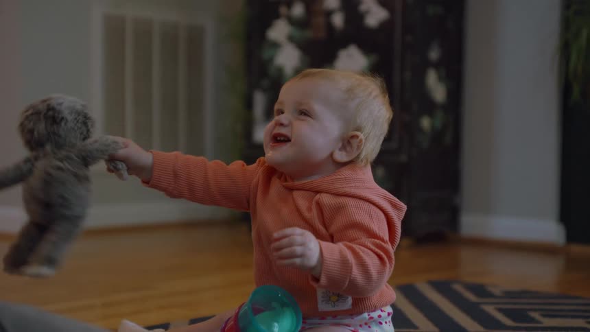 A happy baby in an orange hoodie sits on the floor, plays with a stuffed animal, and then hands it to a parent in slow motion