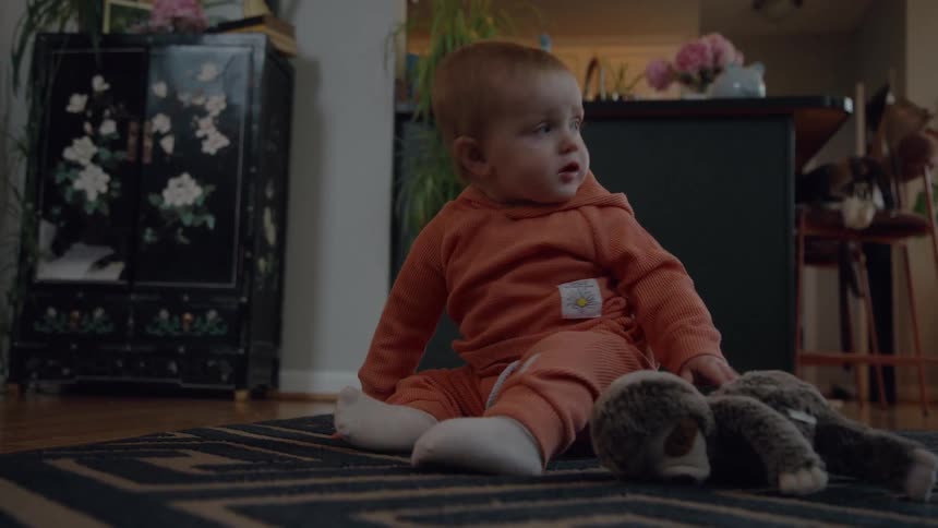 A curious baby in an orange outfit sits on the living room floor playing with a stuffed sloth before looking off-screen