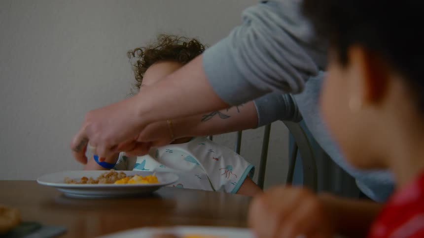 A parent helps their curly-haired toddler eat breakfast by cutting up the food into bite-sized pieces at the dining table