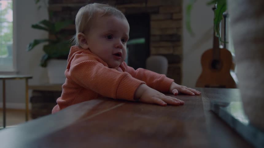 A curious baby in an orange hoodie stands at a wooden table in a living room, waving and patting the tabletop